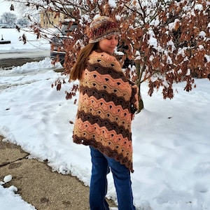 May include: A woman wearing a brown and pink crocheted shawl with a multi-colored crocheted hat. She is standing in a snowy landscape.