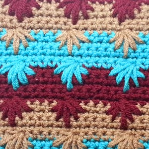 May include: Close-up of a crocheted fabric with a repeating pattern of brown, blue, and maroon leaves. The fabric is made with a variety of stitches and textures.