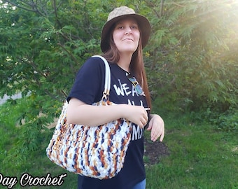 May include: A woman wearing a black t-shirt with the text "We Are All Made" is holding a crocheted tote bag with white, brown, and orange yarn. The bag has a white strap and a gold accent. The woman is standing in front of a green bush.