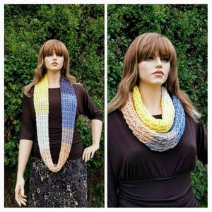 May include: A multi-colored crocheted infinity scarf with shades of yellow, white, peach, and blue. The scarf is worn by a mannequin with long brown hair.