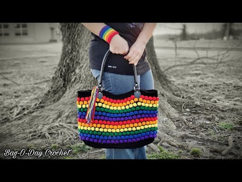 Puede incluir: Un bolso tote de crochet arco&iacute;ris con asas negras y una base negra. El bolso est&aacute; hecho con hilo de colores y tiene un patr&oacute;n de arco&iacute;ris. El bolso lo lleva una persona que lleva una camiseta negra y unos vaqueros azules.