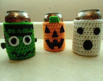 May include: Three crocheted can cozies in green, orange, and white. The green cozy has a Frankenstein face, the orange cozy has a jack-o-lantern face, and the white cozy has a ghost face.