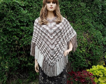 Könnte beinhalten: Ein braunes und weißes gehäkeltes Poncho mit Fransen. Das Poncho hat ein Chevron-Muster und wird von einer Schaufensterpuppe vor einem grünen, bewachsenen Hintergrund getragen.