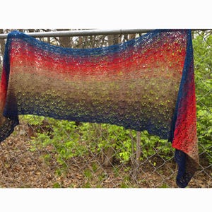 May include: A colourful crocheted shawl with a gradient of red, orange, yellow, green, blue and purple. The shawl is draped over a chain link fence.