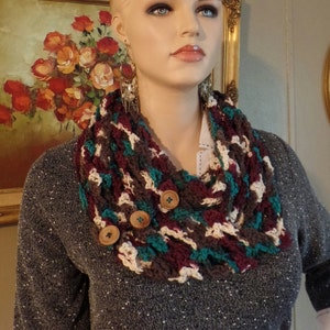 May include: A brown, teal, and white crocheted cowl with brown buttons. The cowl is worn by a mannequin with a gray sweater.