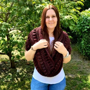 Op de afbeelding: Een vrouw draagt een bordeauxrode cowl-trui met een wit shirt en een blauwe jeans. Ze staat voor een groene, bladerrijke achtergrond.