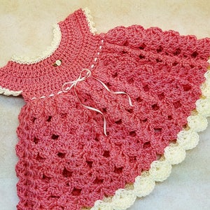 May include: A handmade, pink crochet dress with a scalloped cream-colored trim. The dress features a round neckline, short sleeves, and a delicate white ribbon bow. A small, white flower is sewn near the neckline.