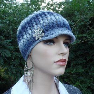 May include: A blue and white crocheted newsboy hat with a snowflake brooch. The hat has a small brim and is made of a soft, textured yarn. The snowflake brooch is silver and adds a touch of sparkle. The hat is a winter accessory.