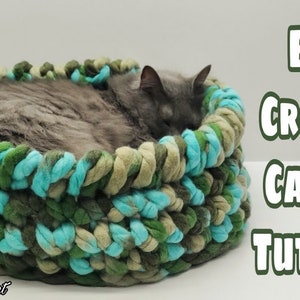May include: A grey cat sleeping in a crocheted cat bed made with green, blue and beige yarn. The bed is round and has a raised edge. The text "EASY CROCHET CAT BED TUTORIAL" is written in white letters on a white background.