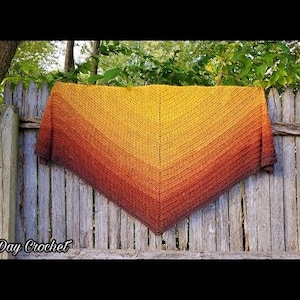 May include: A crocheted shawl with a gradient of orange, yellow, and brown colors. The shawl is draped over a wooden fence.