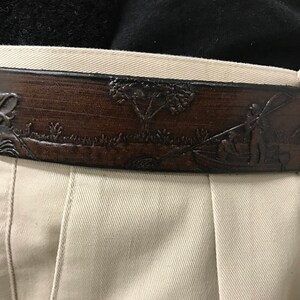 May include: A brown leather belt with an embossed design of a fish, a tree, and a person fishing. The belt is worn on a pair of beige pants.