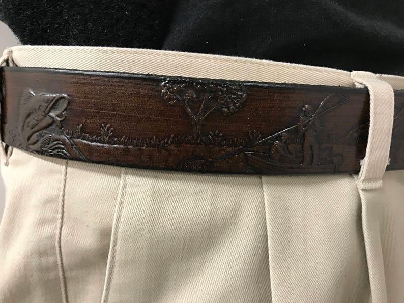 May include: A brown leather belt with an embossed design of a fish, a tree, and a person fishing. The belt is worn on a pair of beige pants.