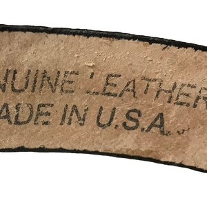May include: Close-up of a brown leather belt with black stitching. The belt has a label that reads "GENUINE LEATHER MADE IN U.S.A."