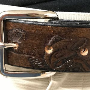 May include: A brown leather belt with a silver buckle. The belt has an embossed design of a fish.