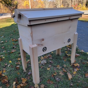 May include: A wooden beehive with a light-colored finish, featuring a sloped roof and four sturdy legs. The hive has two circular vents on the front and is set on a grassy area with fallen leaves.