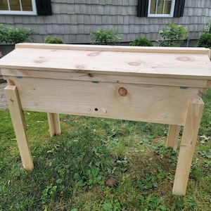 May include: A wooden box with a hinged lid and four legs. The box is made of unfinished pine wood and has a natural wood grain. The box is sitting on a bed of green grass.