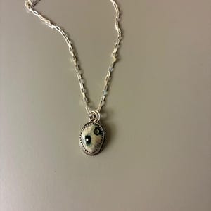 May include: A silver necklace with a small, oval pendant. The pendant has a white and blue design with a silver border. The chain is a delicate, linked style. The necklace is displayed against a gray background.