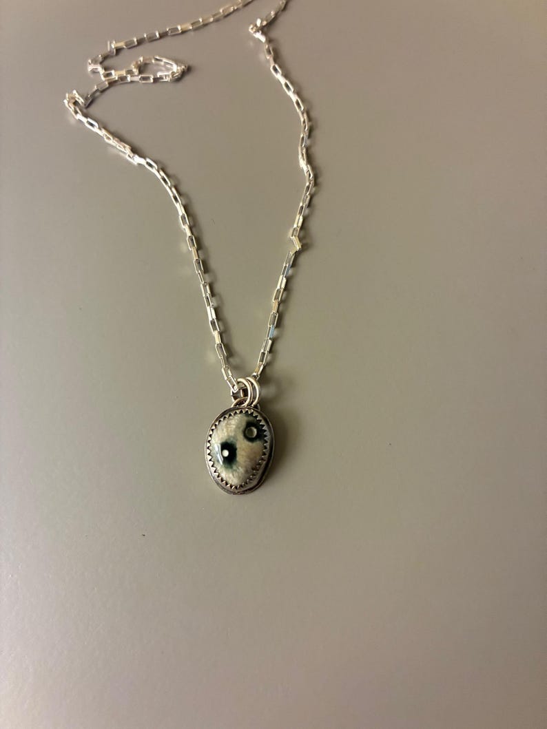 May include: A silver necklace with a small, oval pendant. The pendant has a white and blue design with a silver border. The chain is a delicate, linked style. The necklace is displayed against a gray background.