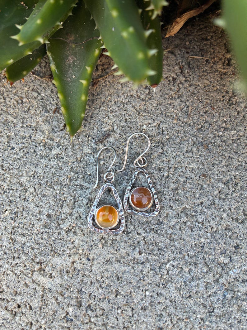 Puede incluir: Un par de pendientes de plata con dise&ntilde;o de l&aacute;grima. Cada pendiente presenta un marco plateado texturizado que rodea una piedra redonda de color &aacute;mbar. Los pendientes se muestran sobre una superficie gris texturizada.