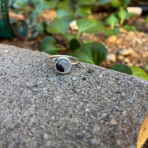 May include: A silver ring featuring a circular, two-tone black and white stone. The ring sits on a grey, textured surface, with green foliage in the background. The ring's band is slender and delicate.