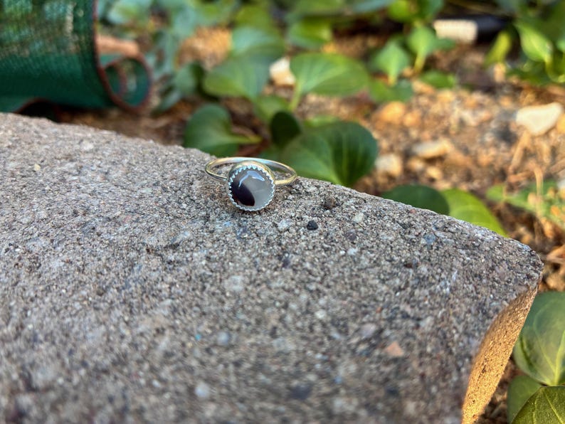 May include: A silver ring featuring a circular, two-tone black and white stone. The ring sits on a grey, textured surface, with green foliage in the background. The ring's band is slender and delicate.