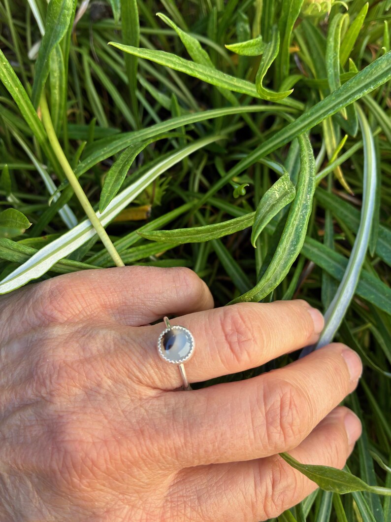 May include: A silver ring featuring a round, pale blue and grey stone. The ring has a simple, minimalist design. The ring is displayed on a hand, with green foliage visible in the background.