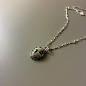 May include: A silver necklace featuring a small, oval pendant. The pendant has a light-colored background with dark, organic-shaped details. The pendant is framed by a textured silver border and hangs from a delicate silver chain.