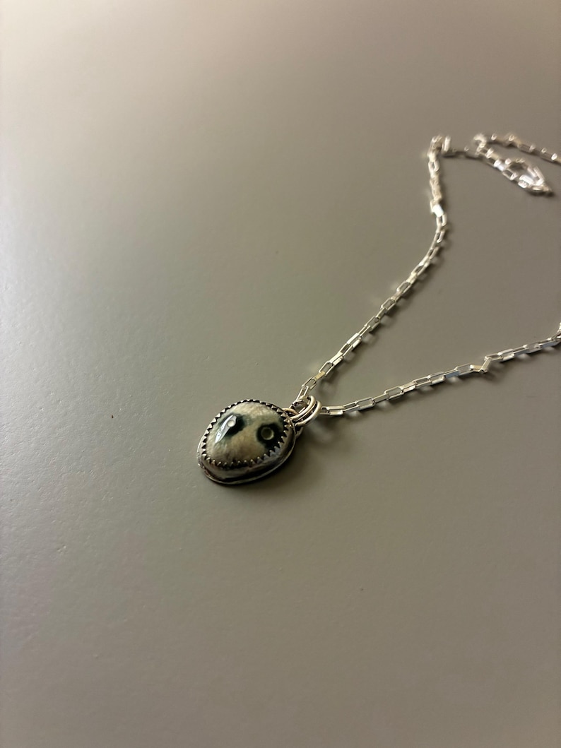 May include: A silver necklace featuring a small, oval pendant. The pendant has a light-colored background with dark, organic-shaped details. The pendant is framed by a textured silver border and hangs from a delicate silver chain.