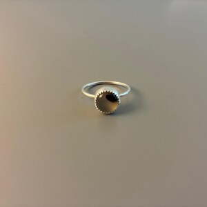 May include: A silver ring featuring a circular, dark grey stone. The stone is set within a bezel with a scalloped border. The ring rests on a grey surface. This is a piece of jewellery.