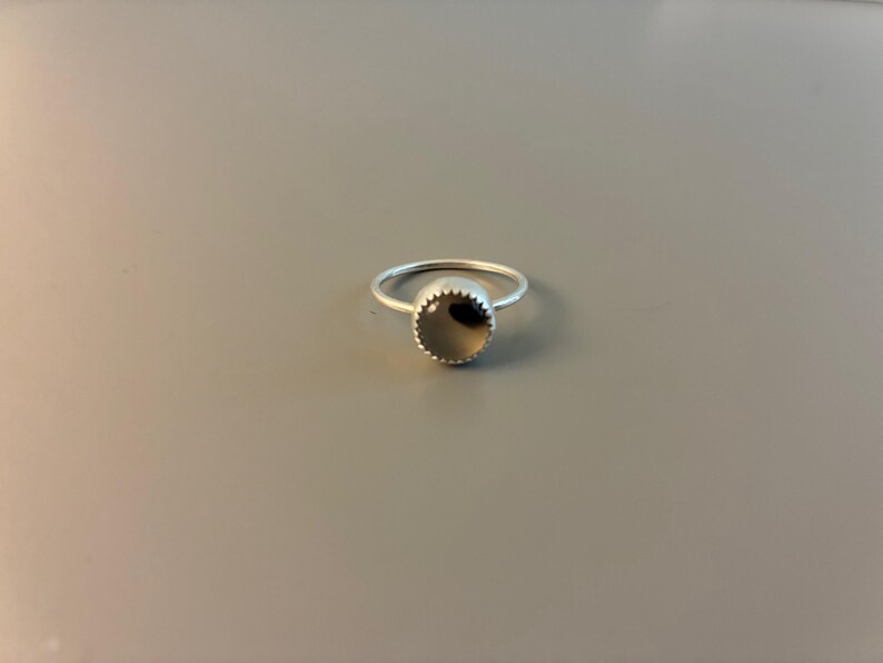May include: A silver ring featuring a circular, dark grey stone. The stone is set within a bezel with a scalloped border. The ring rests on a grey surface. This is a piece of jewellery.
