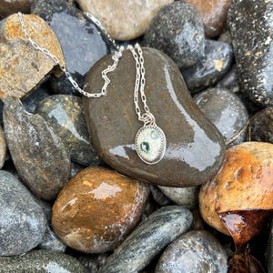 May include: A silver necklace with an oval pendant featuring a light green and white design. The pendant hangs on a delicate chain, resting on a collection of wet, smooth stones in various shades of brown, gray, and tan.
