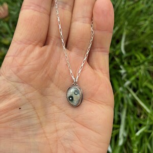May include: A silver necklace featuring a small, oval pendant. The pendant has a light-colored background with two dark green spots and a textured, silver border. The chain is a delicate, linked design.