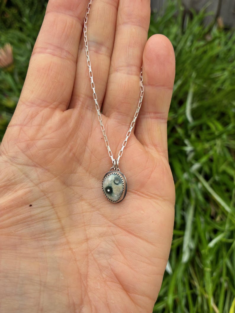 May include: A silver necklace featuring a small, oval pendant. The pendant has a light-colored background with two dark green spots and a textured, silver border. The chain is a delicate, linked design.