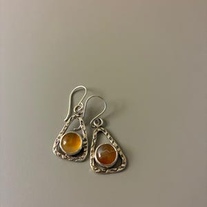 Puede incluir: Un par de pendientes de plata con un marco texturizado y forma de l&aacute;grima. Cada pendiente presenta una piedra redonda de color &aacute;mbar. Los pendientes tienen un cierre de gancho y se muestran sobre un fondo gris.