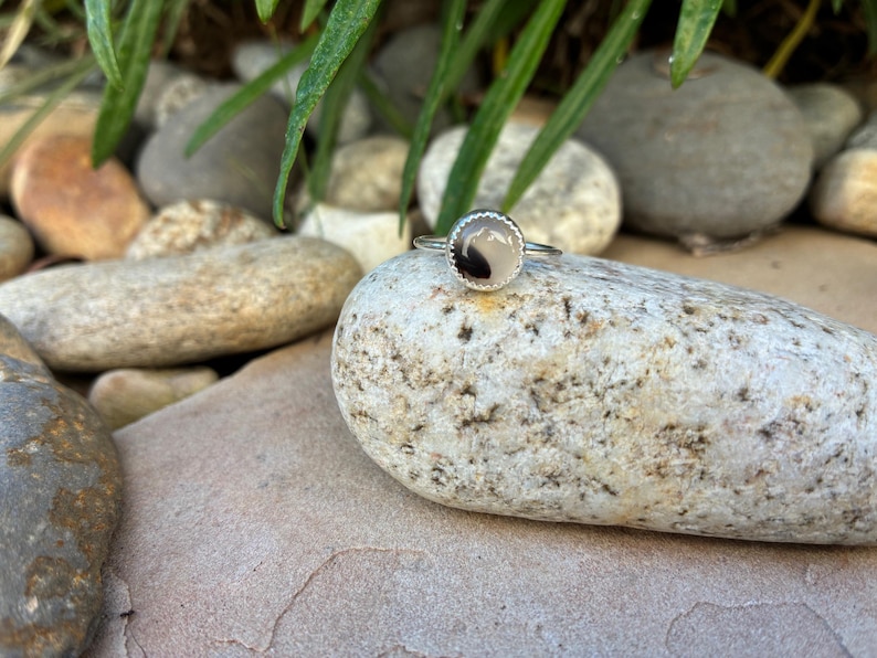 May include: A silver ring featuring a circular stone with a light base and a dark, organic design. The ring is set on a light-coloured rock, with other rocks and foliage in the background. This ring is a piece of jewellery.