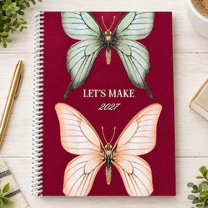 May include: A spiral-bound notebook with a deep red cover featuring two large butterflies. The top butterfly is pale green, and the bottom butterfly is light orange. The text "LET'S MAKE 2027" is printed in white in the center between the butterflies.