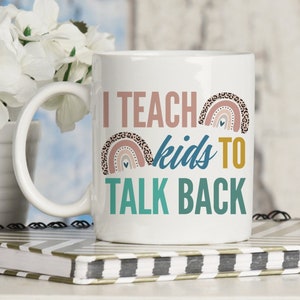 Puede incluir: Taza de café de cerámica blanca con un diseño en azul verdoso, rosa y marrón. La taza dice "Enseño a los niños a responder".