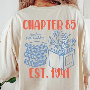 Puede incluir: Camiseta color marfil con el texto "CHAPTER 85" en coral. Debajo, se lee "Nicole's 85th birthday" y "EST. 1941". El diseño incluye ilustraciones de libros azules y detalles florales. La camiseta es Comfort Colors 1717, talla L.