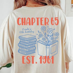 Pode incluir: Camiseta marfim com o texto "CHAPTER 65" em coral. Abaixo, lê-se "Nicole's 65th birthday" e "EST. 1961". O design inclui livros empilhados e um livro com flores. A camiseta é Comfort Colors 1717, tamanho L.