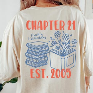 May include: Ivory-colored t-shirt with the text "CHAPTER 21" in coral. Below, it reads "Nicole's 21st birthday" and "EST. 2005." The design includes blue book illustrations and floral elements. The shirt is a Comfort Colors 1717.