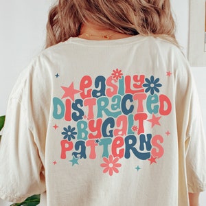 Puede incluir: Una camiseta de color crema con un estampado gráfico que dice "easily distracted by patterns" en una fuente retro con colores rosa, azul y verde.
