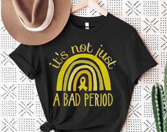 Puede incluir: Camiseta negra con un gráfico de arcoíris amarillo y el texto "it's not just a bad period".
