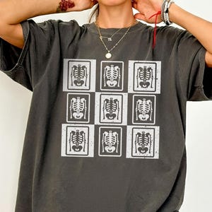 May include: Dark grey t-shirt with a grid of nine white skeleton illustrations. The shirt is a Comfort Colors brand in the color Pepper. The model is wearing an XL size.
