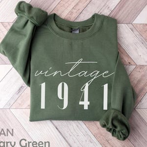 Op de afbeelding: Militairgroene sweatshirt met de tekst "vintage 1941" in witte letters. De sweatshirt heeft een ronde hals en lange mouwen. De merknaam "GILDAN" en de kleur "Military Green" staan onderaan gedrukt. De stof lijkt zacht.