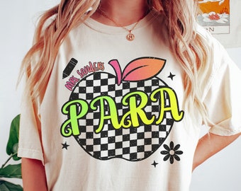 Könnte beinhalten: Ein weißes T-Shirt mit einem schwarz-weiß karierten Apfel-Design. Der Apfel hat einen grünen Stiel und rosa und orangefarbene Blätter. Der Text "Mrs. Sanders PARA" ist in einer bunten Schrift auf dem Apfel geschrieben.