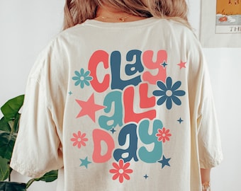 May include: A cream colored t-shirt with a retro-style graphic that says "Clay All Day" in pink, blue, and teal with stars and flowers. The model is wearing a size large.