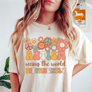 May include: A cream colored t-shirt with a retro-style graphic featuring flowers, peace signs, and the text "AUTISM seeing the world DIFFERENTLY".