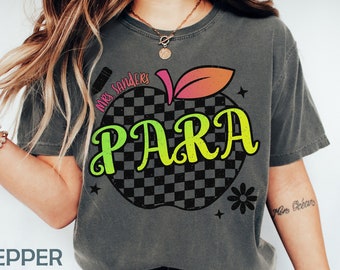 Könnte beinhalten: Ein graues T-Shirt mit einem schwarz-weiß karierten Apfeldesign. Der Apfel hat einen grünen Stiel und ein Blatt und das Wort "PARA" in grünen und rosa Buchstaben. Der Text "Mrs. Sanders" ist in Schwarz auf den Apfel geschrieben.