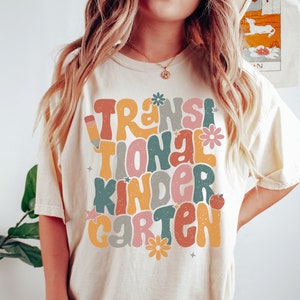 May include: Ivory t-shirt with the text "Transitional Kindergarten" in a retro font with colorful letters and flowers. The model is wearing a size large.