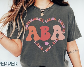 Könnte beinhalten: Ein graues T-Shirt mit den Buchstaben "ABA" in Rosa, Orange und Rot. Die Buchstaben sind von einem Kreis mit den Worten "Compassionate, Caring, Dedicated, Reliable, Loyal, Kind" umgeben.